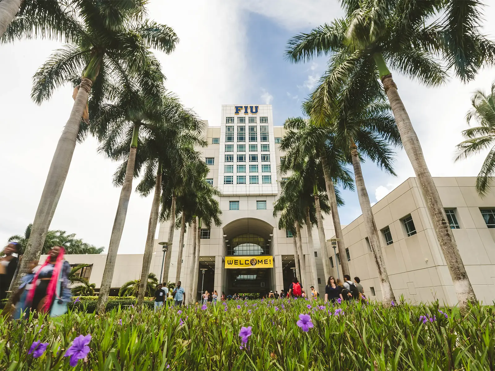 View of FIU campus with tall palm trees lining a walkway, students walking, and the main building with a 'Welcome!' sign in the background.