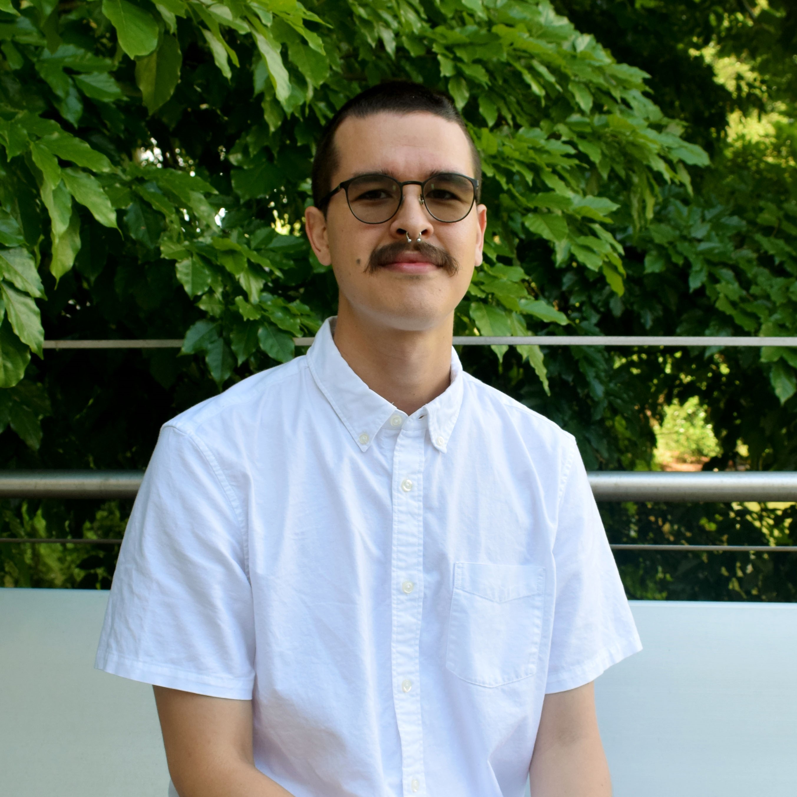 A young, mustached man wearing lightly-tinted black sunglasses and a white shirt poses outdoors for a professional photo.