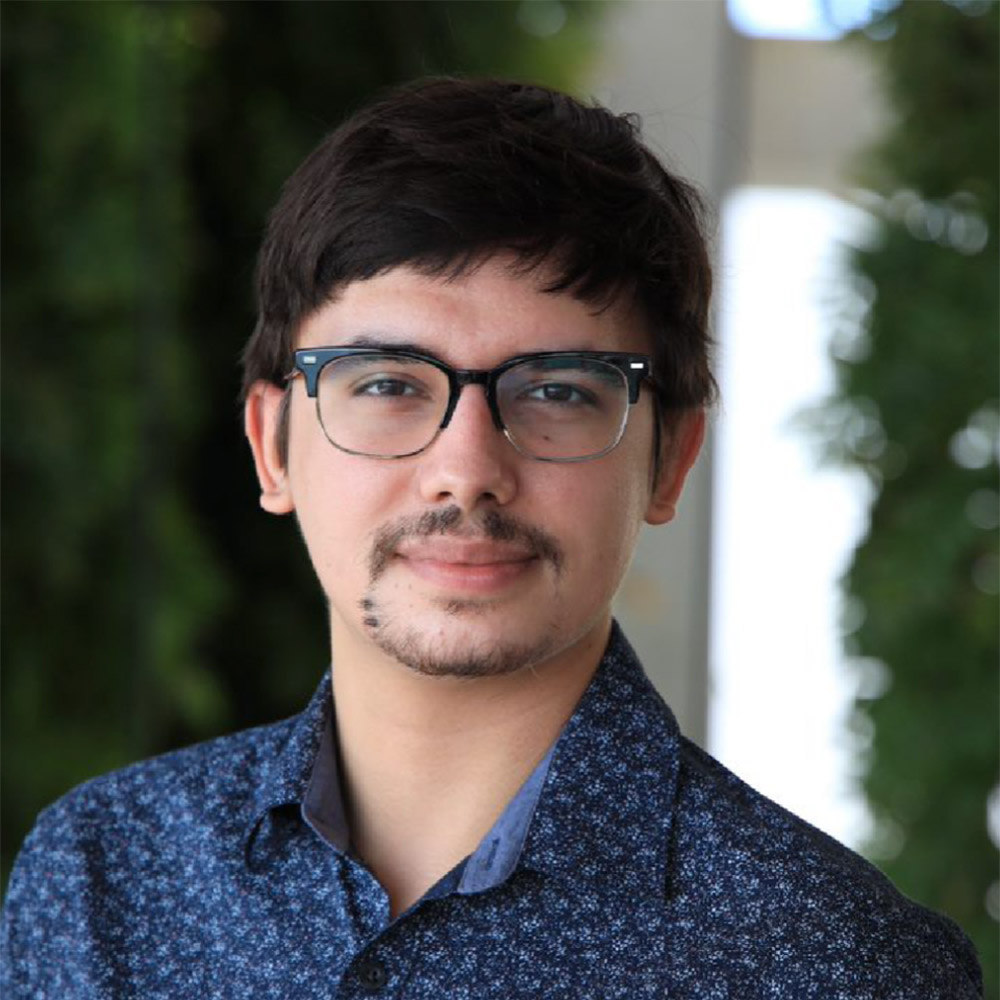 A young man with short dark hair, thin-rimmed black glasses, and a small mustache and goatee poses for a professional outdoor headshot.