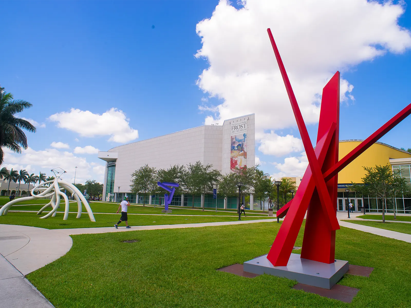 View of the Avenue of the Arts at FIU, featuring a red abstract sculpture and the Frost Art Museum in the background. Other art installations are visible around the area.