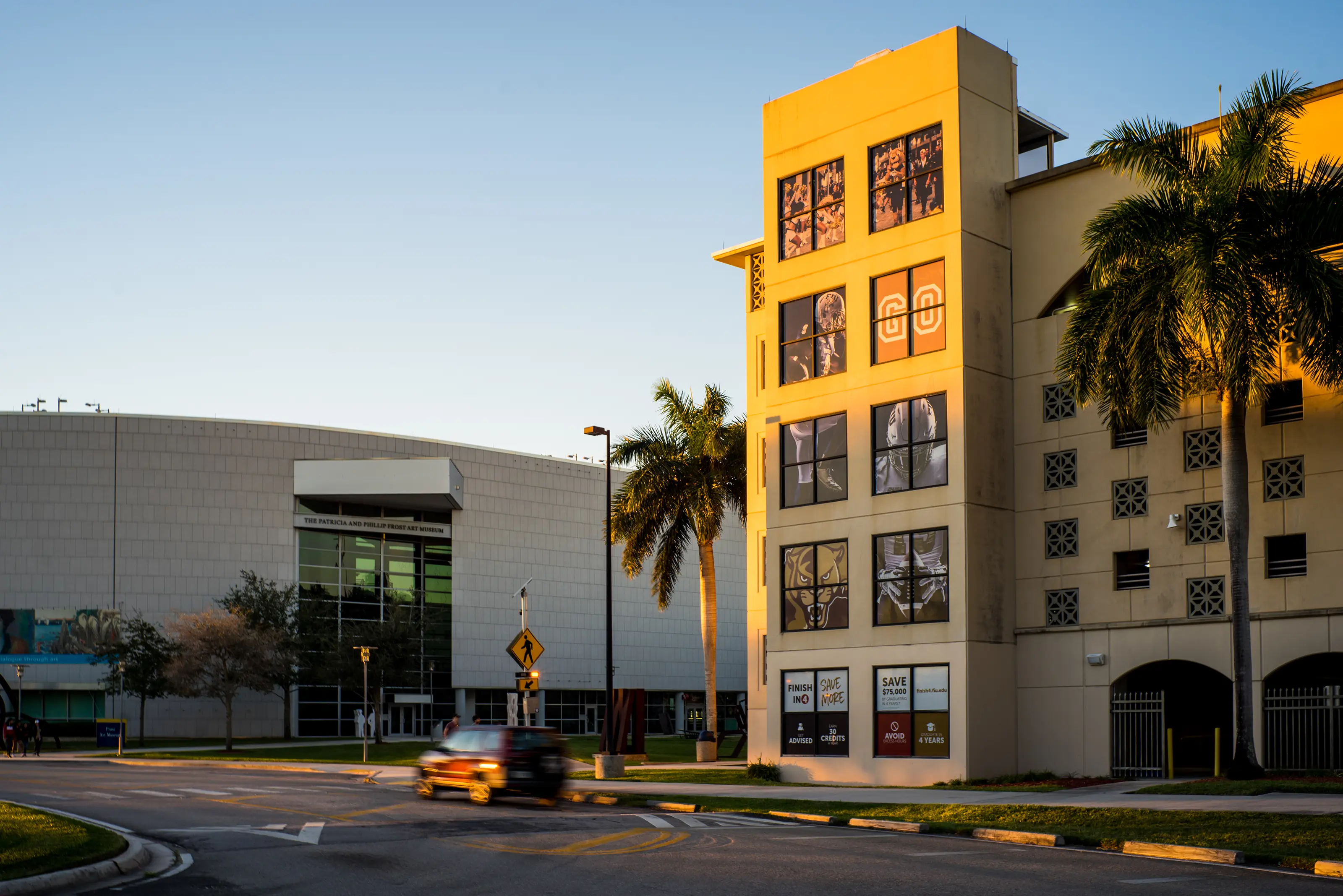 Exterior view of the Blue Garage with decorative panels next to the Frost Art Museum at Florida International University during golden hour, with a blurred vehicle passing by on the street in front.